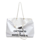Angled front view of the Caffeinated & Investigating Weekender Bag displaying bold investigator-themed typography and magnifying glass design on white spun polyester