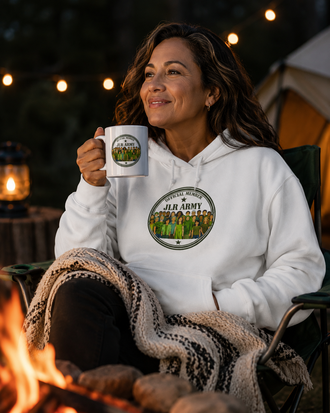 woman drinking coffee by campfire wearing White premium hoodie featuring the JLR© Army circular badge graphic with diverse illustrated figures on the chest