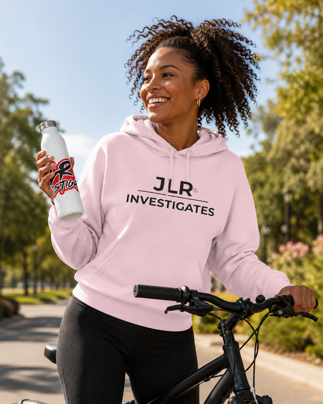 Woman on bicycle wearing light pink pullover hoodie with JLR Investigates logo in black