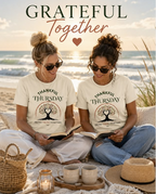 Two women sitting on beach wearing cream Thankful Thursday t-shirts with tree of life graphic, Grateful Together lifestyle photo